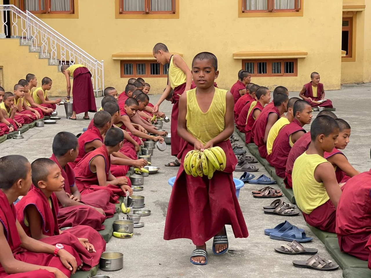 A child carrying bananas between rows of children seated for a meal.