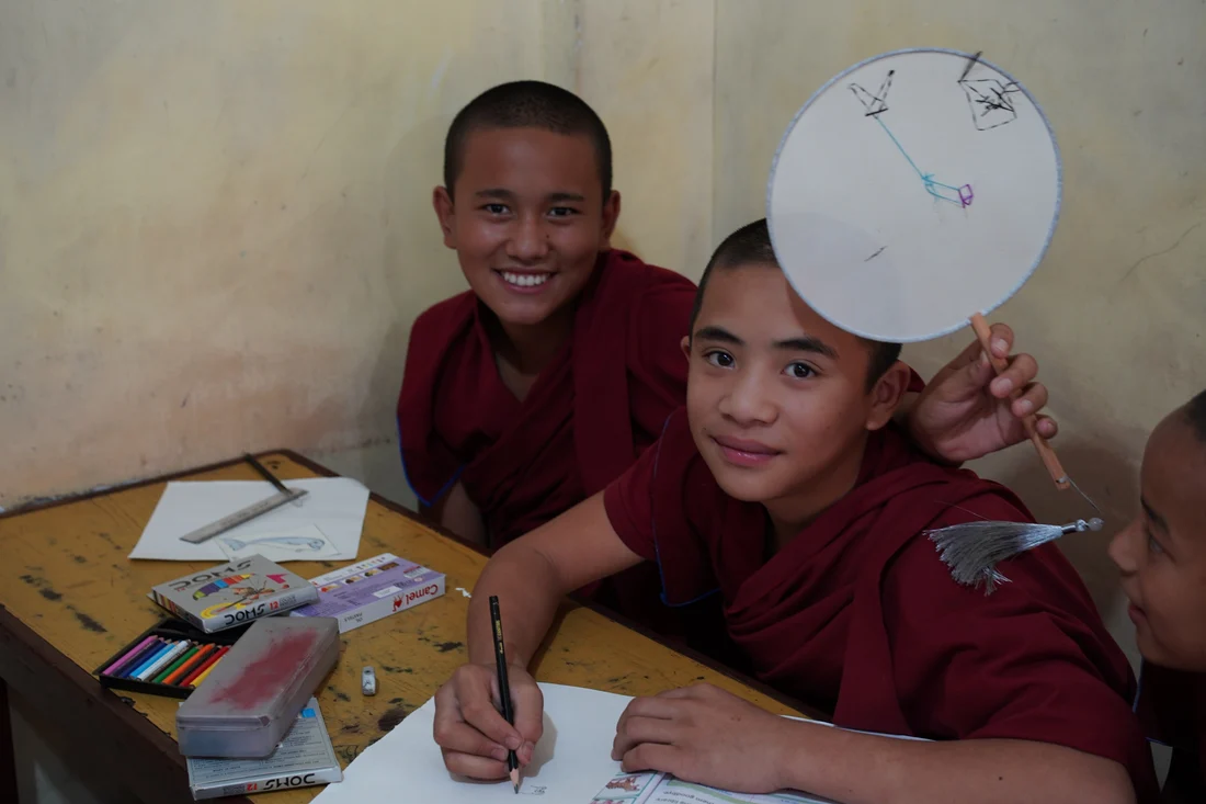 Children at a classroom desk drawing, one holding up a round fan with a tassel.